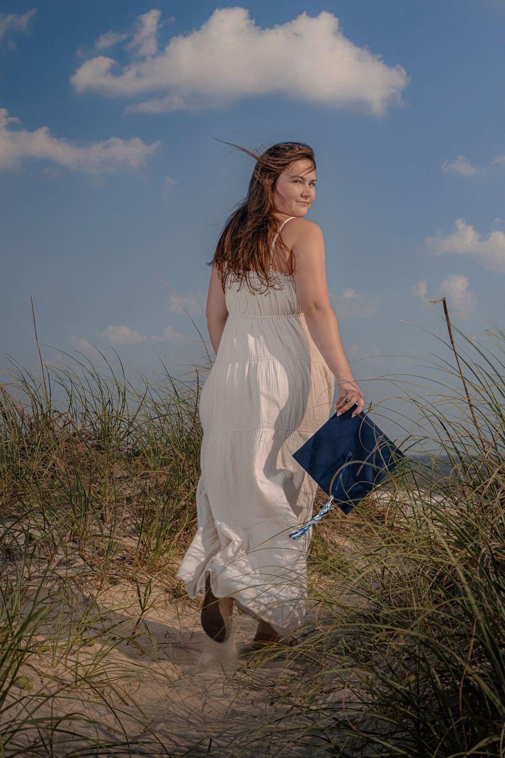 A person in a white dress sits among beach grass on a sand dune against a blue summer sky.