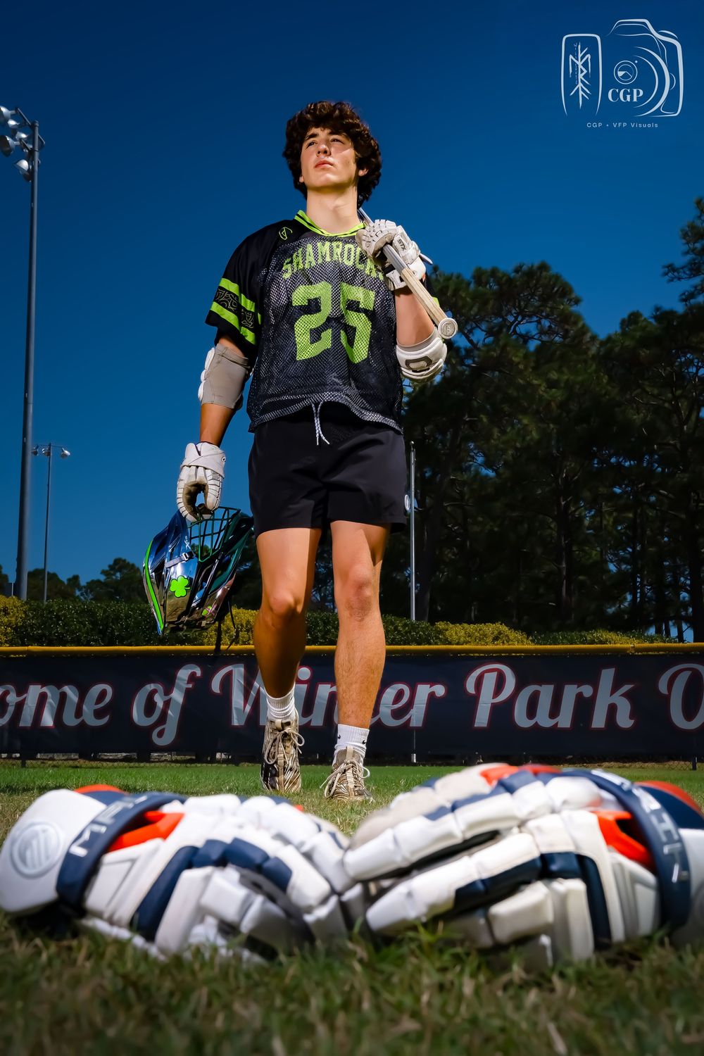 Soccer cleats and equipment lay on a field while a player stands in the background on a sunny day.