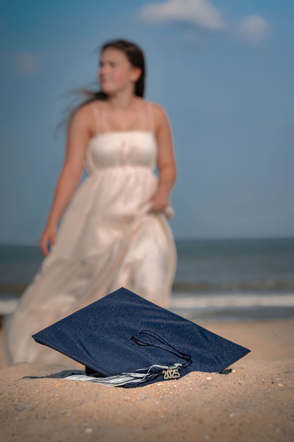 A graduation cap rests on sandy beach with someone in a white dress standing in the blurred background.