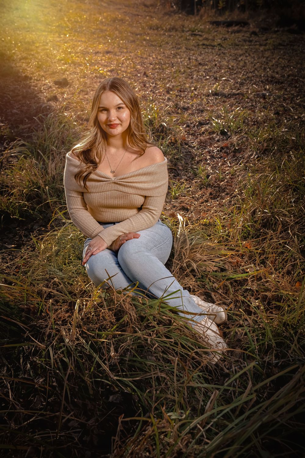 Person in beige sweater and jeans sits on autumn ground during golden hour photoshoot in nature.