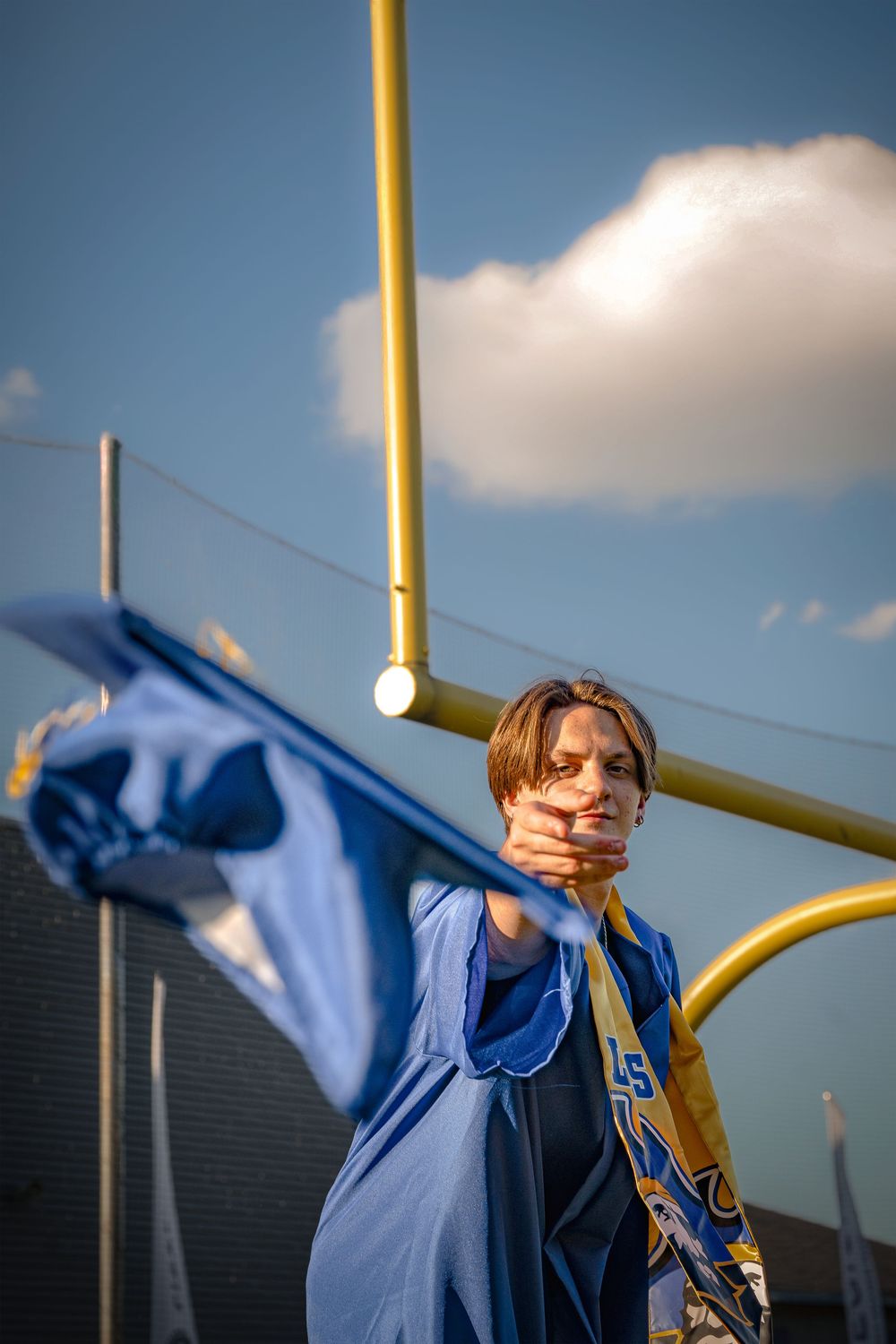 A person in a blue jacket stands against a cloudy sky with yellow goal posts visible in the background.