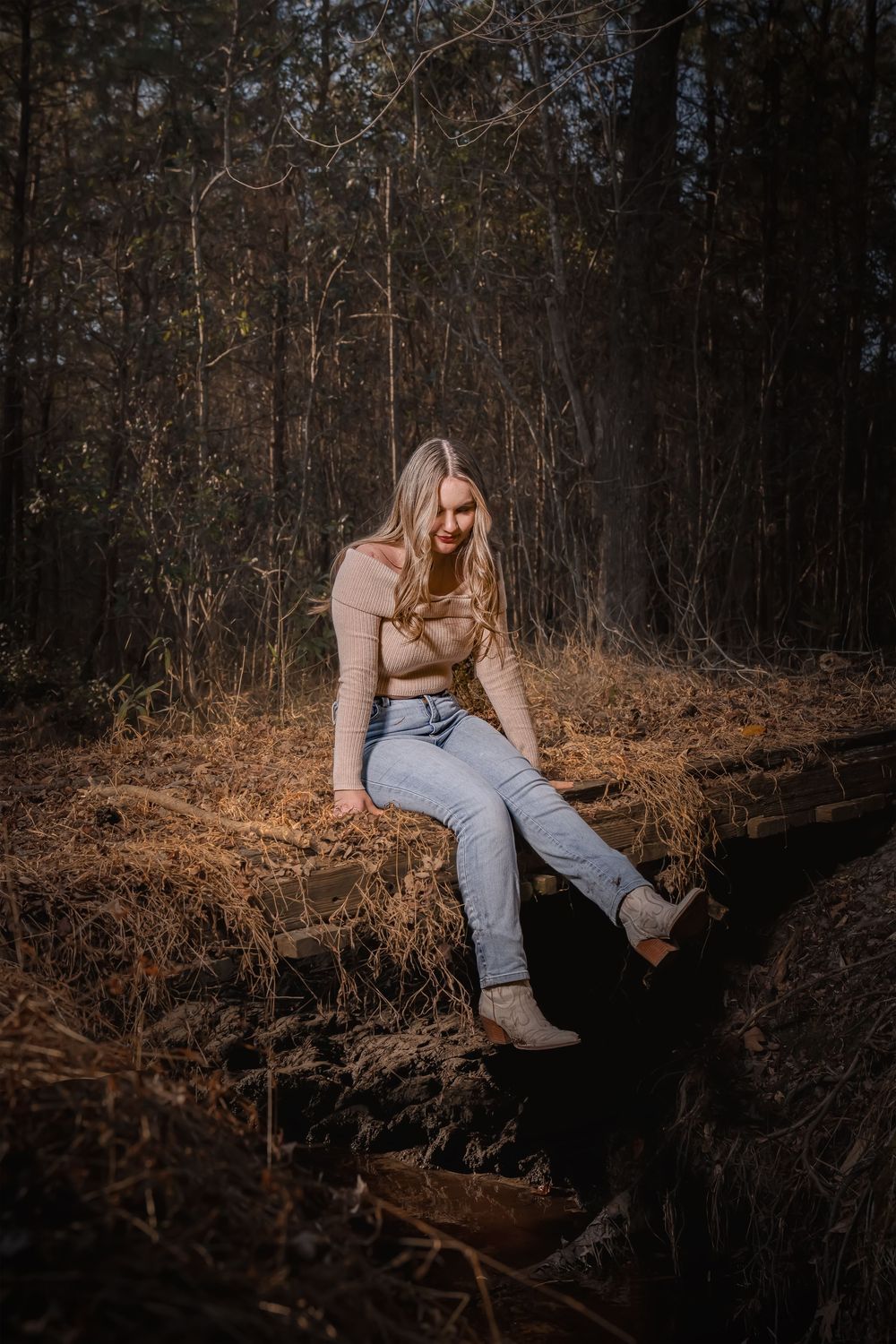 Person in beige sweater and jeans sits on fallen log in autumn forest during golden hour.