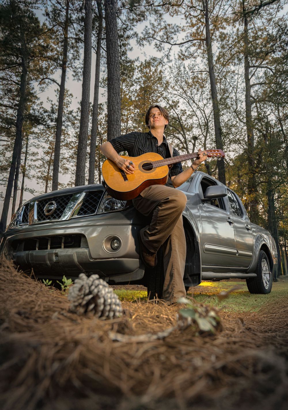 A musician sits on the hood of a silver car in the woods playing acoustic guitar during an autumn day.