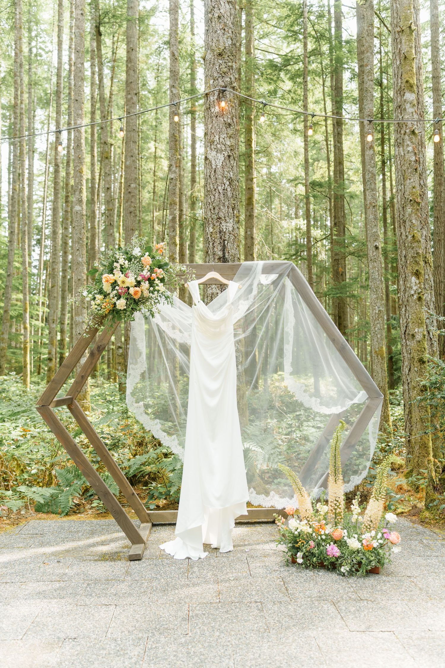 A white wedding dress displayed on a geometric wooden arch with floral arrangements in a serene forest setting.