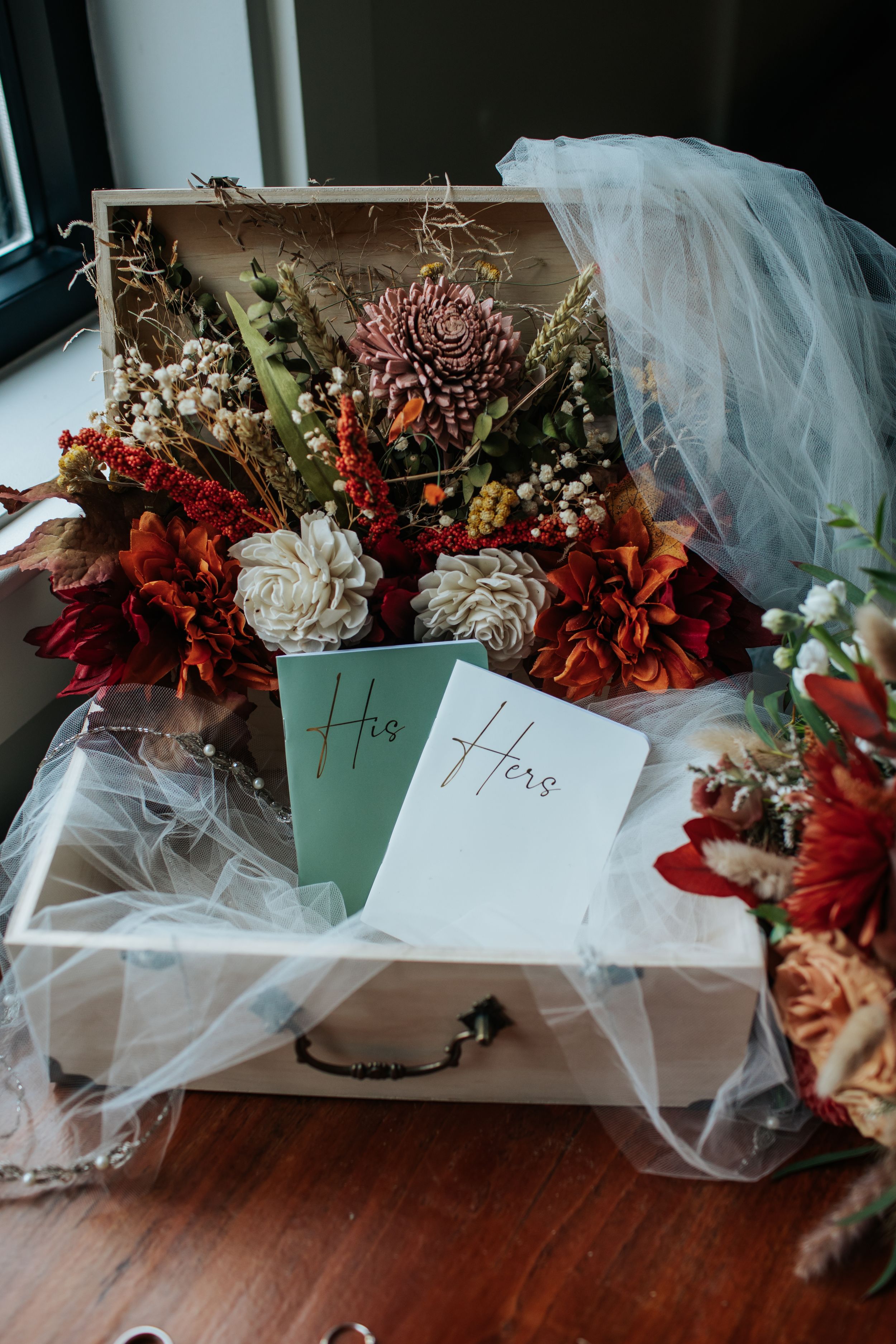 Wedding bouquets wrapped in plastic with name tags sit on a wooden surface, featuring red and burgundy flowers.