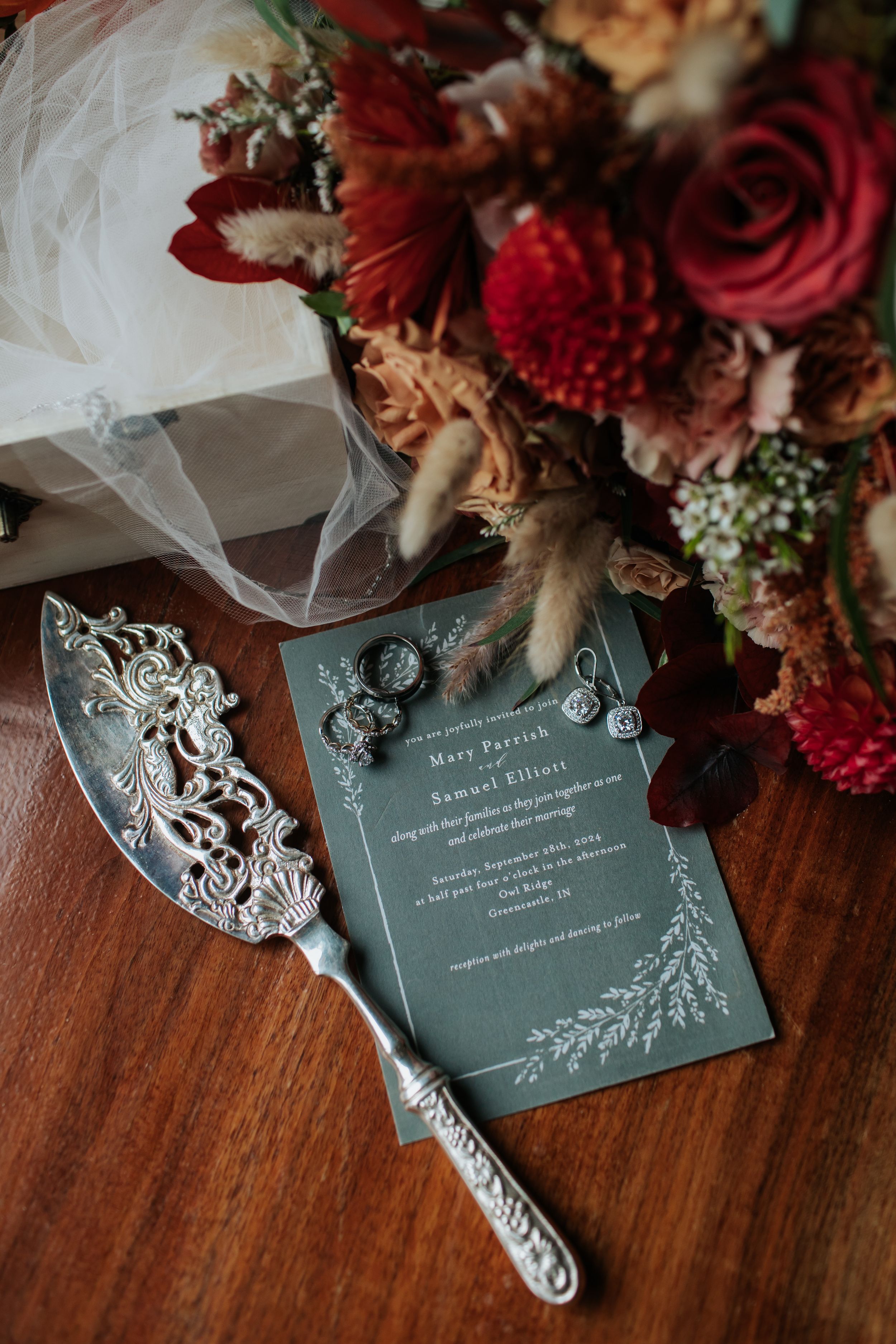 Wedding invitation and details card in sage green with elegant floral designs lay beside red and burgundy flowers on wooden table.