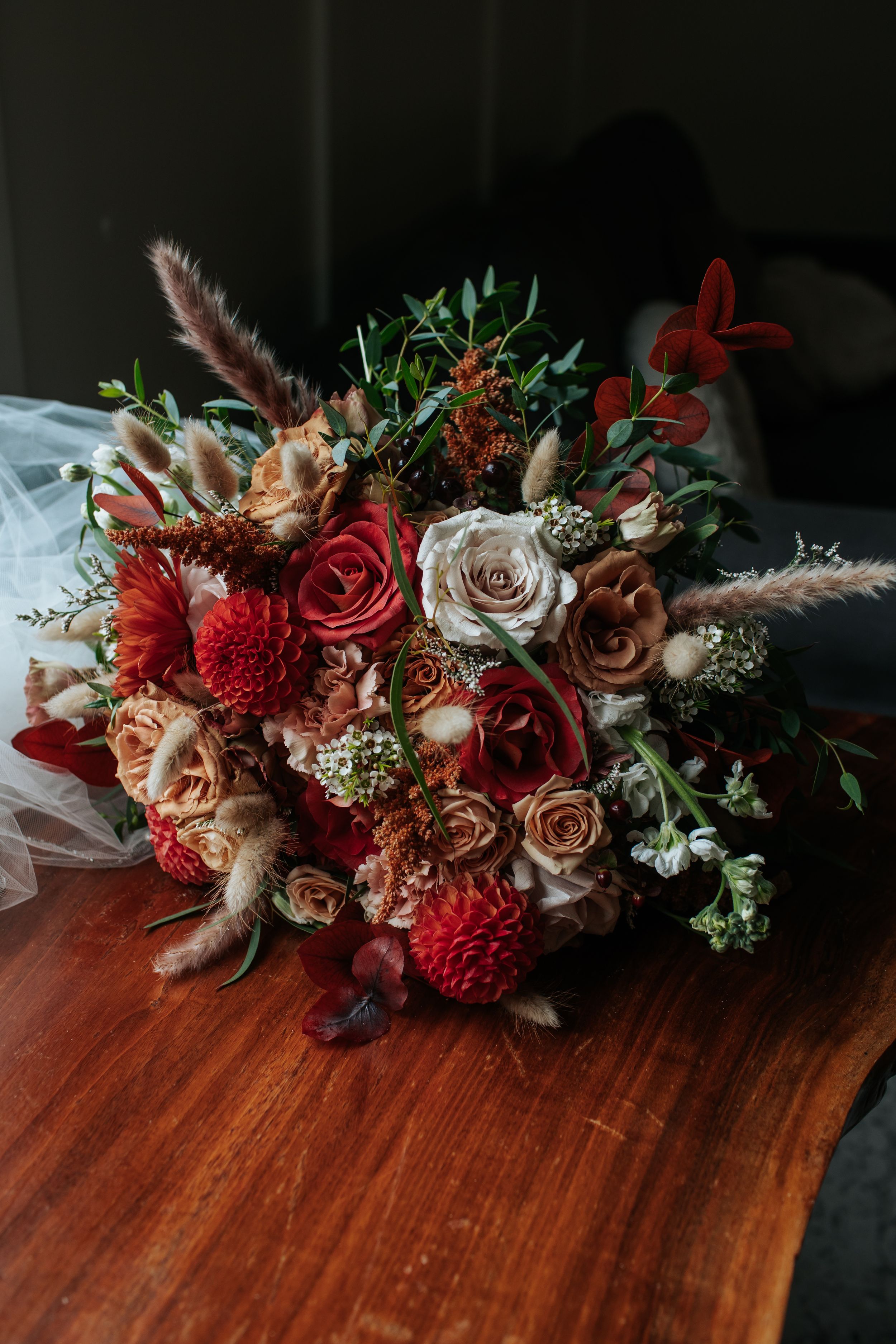 Romantic floral arrangement with deep red roses, white blooms and dried elements on a dark wooden surface.