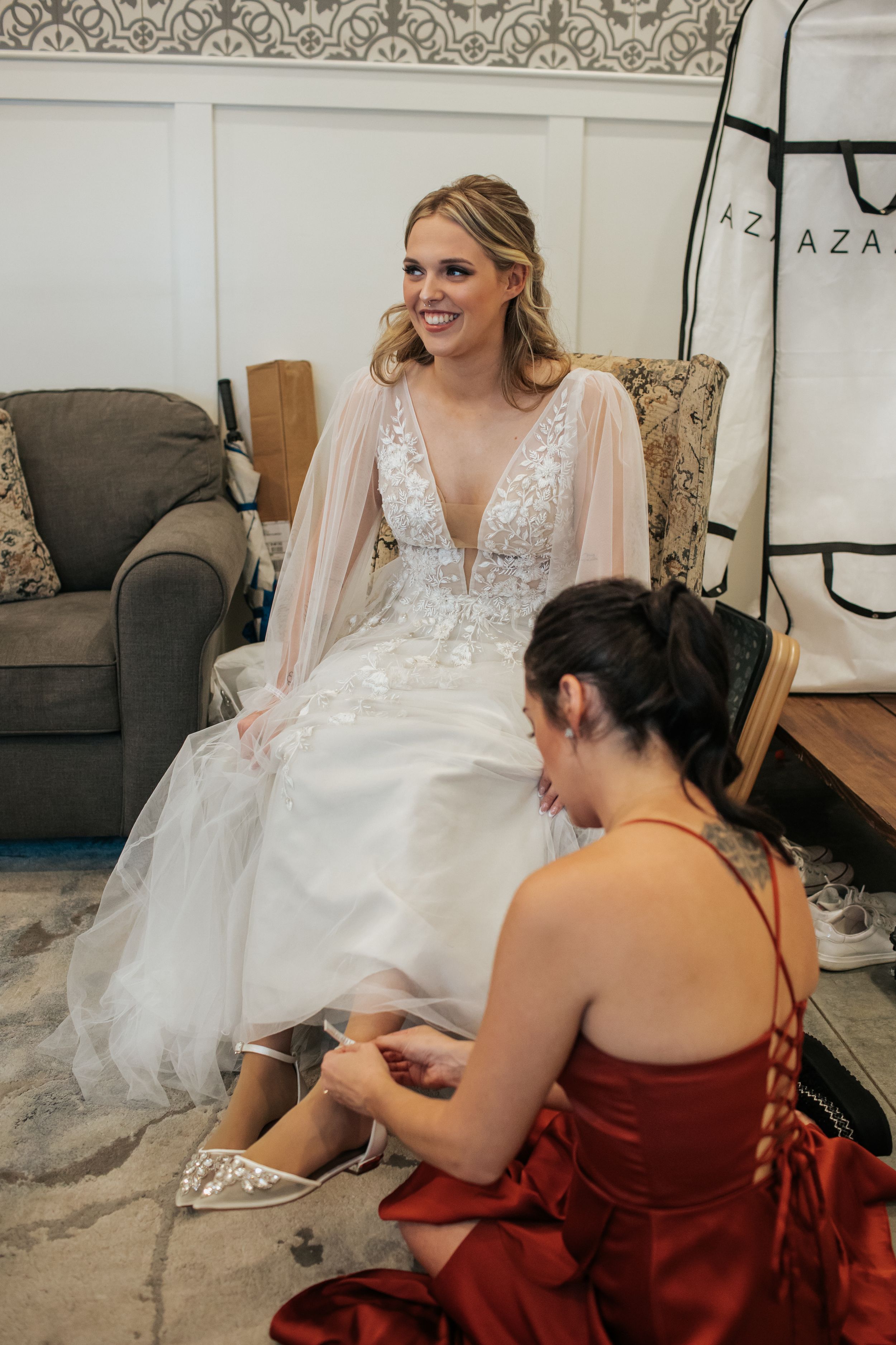 A bride in a flowing white gown sits while someone in burgundy adjusts her dress during wedding preparations.