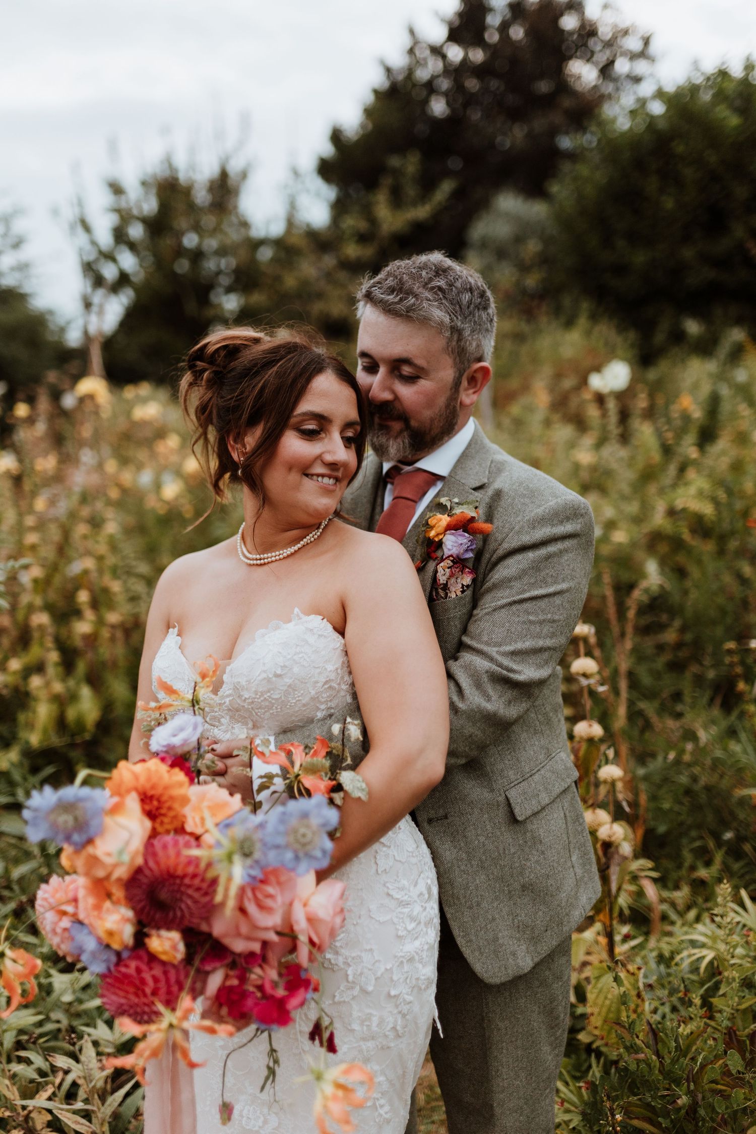 Romantic wedding couple embraces in a wildflower meadow with colorful summer bouquet at golden hour.