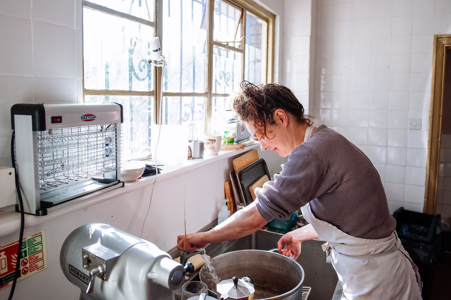 Person working in a bright commercial kitchen with white tiled walls and large windows.