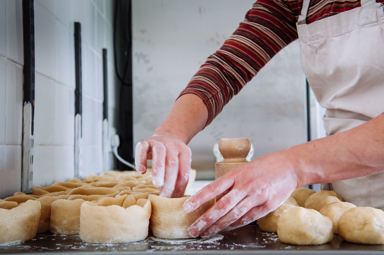Hands rolling and shaping fresh bread dough on a floured wooden surface in a bakery kitchen.