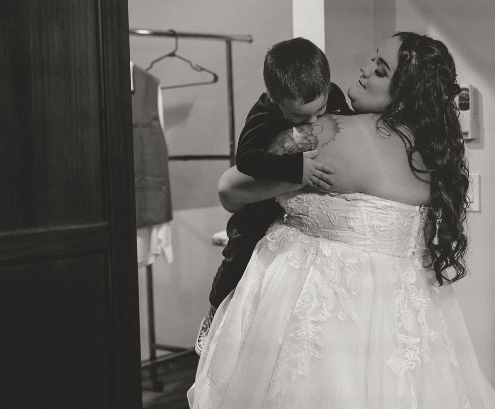 Black and white photo of a bride in a white gown embracing a young boy in a suit on her wedding day.