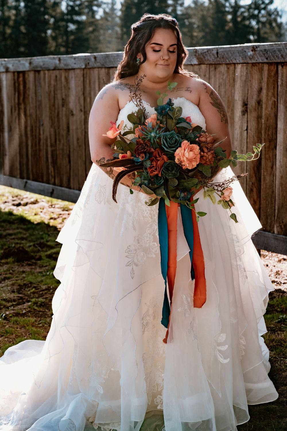 Plus-size bride in layered white gown holding lush bouquet with teal and orange ribbons, antlers, and succulents.