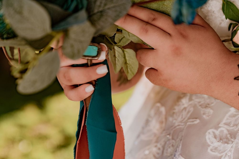 Close-up of bride holding bouquet with turquoise ring visible, teal and orange ribbon streamers, lush greenery.