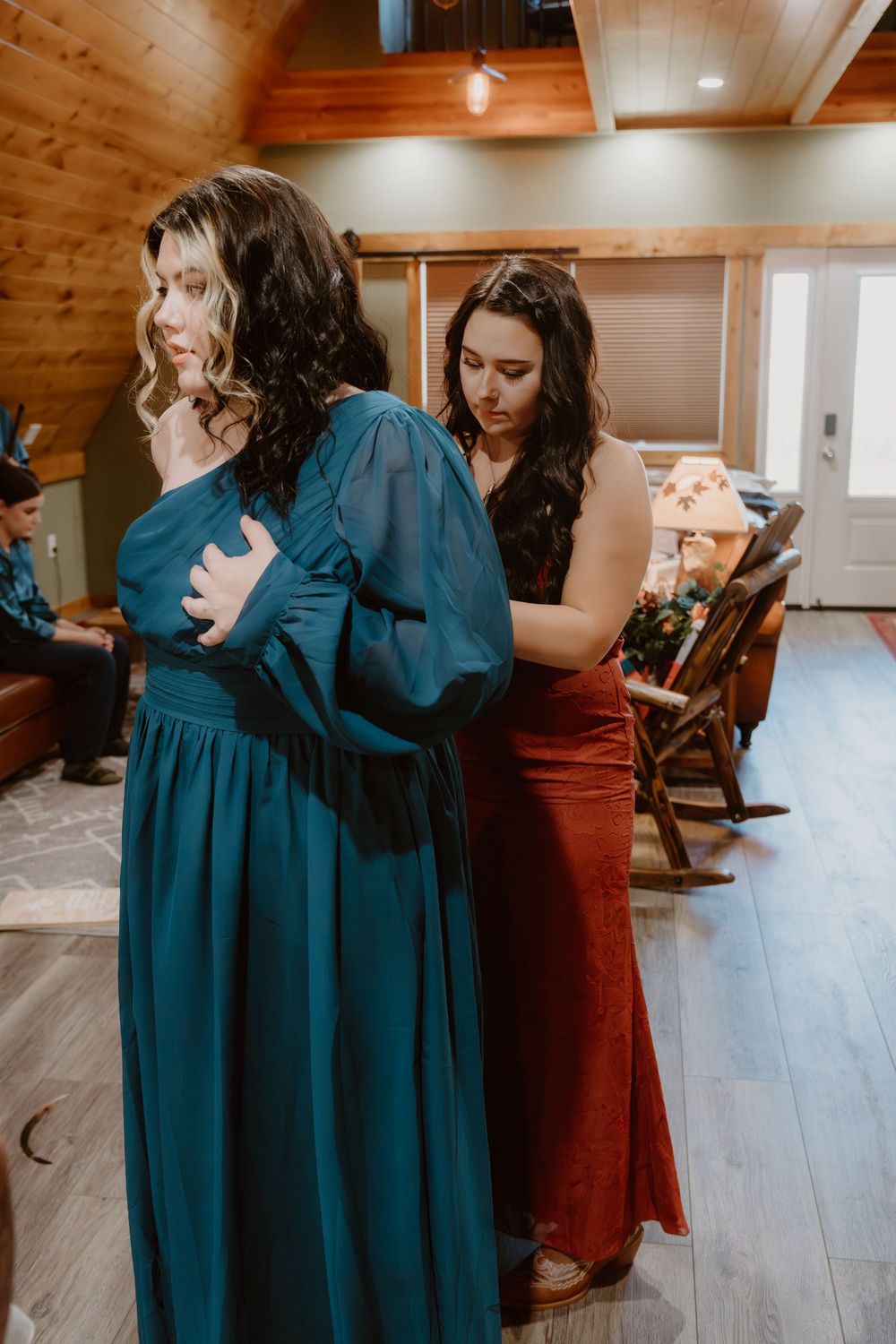 Two women in elegant teal and rust gowns stand in a rustic cabin during a wedding preparation scene.