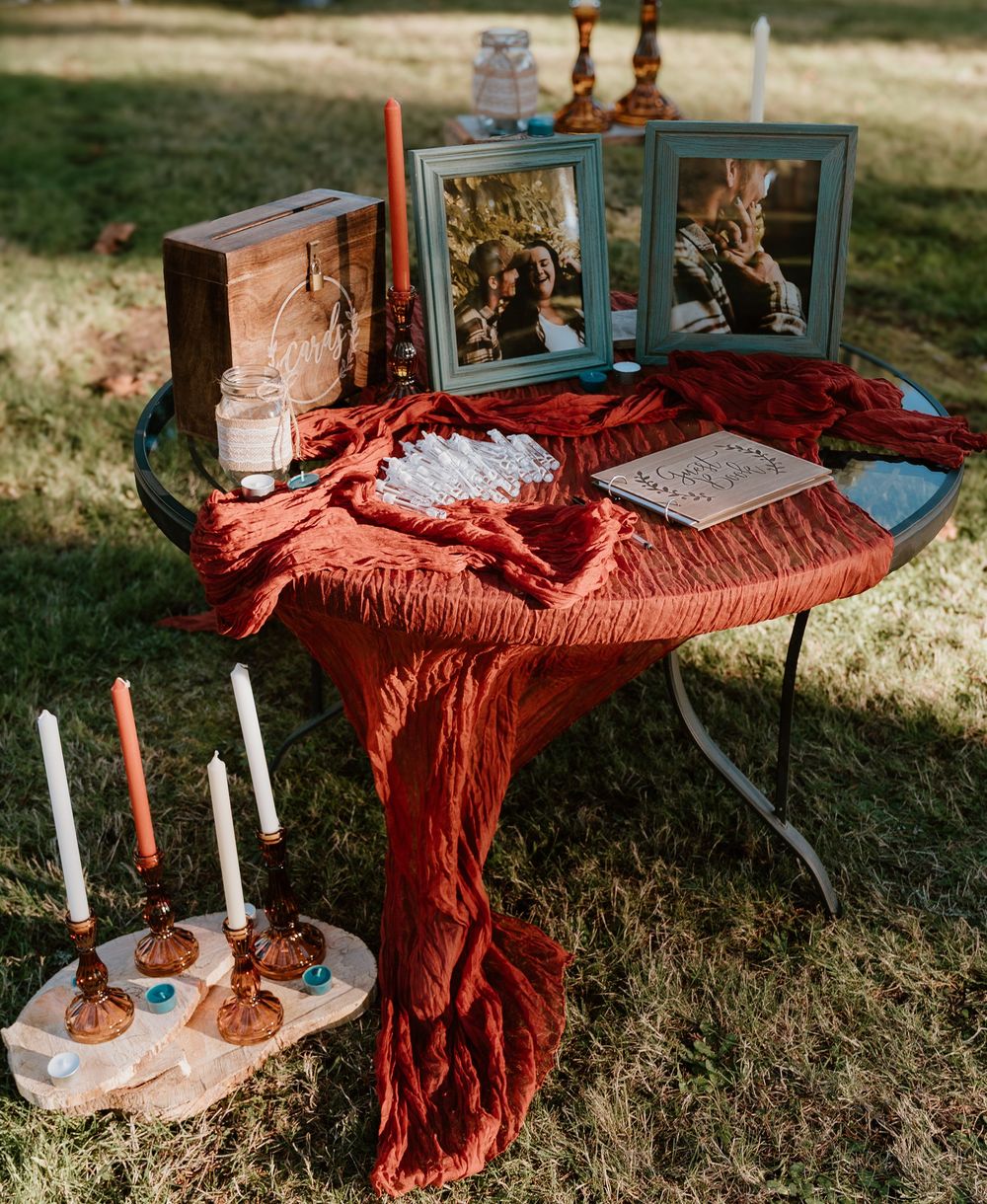 Rustic outdoor wedding table with red draped cloth, framed photos, candles, and a guest book on green grass.
