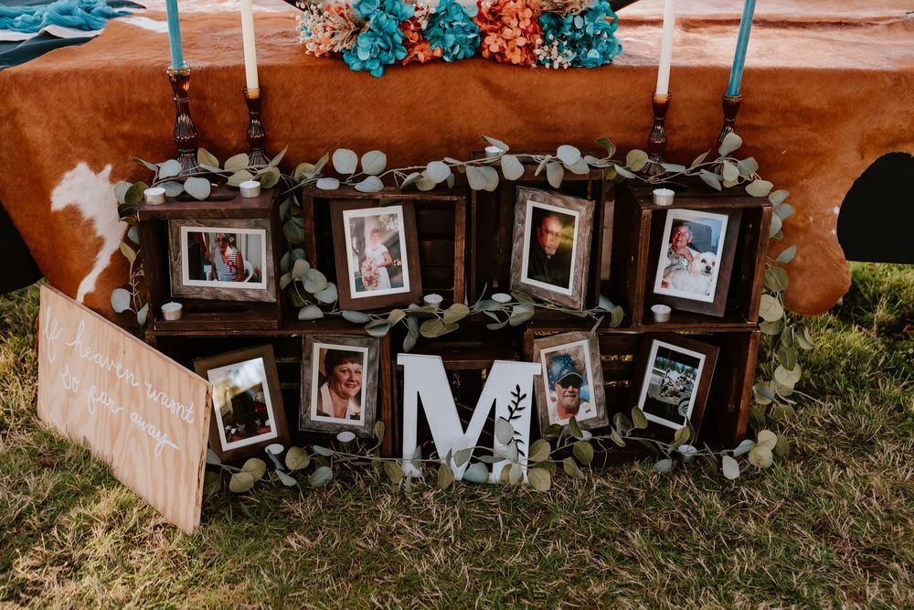Rustic wedding memorial display with framed photos arranged on wooden crates, eucalyptus garland, and decorative letter M on grass.