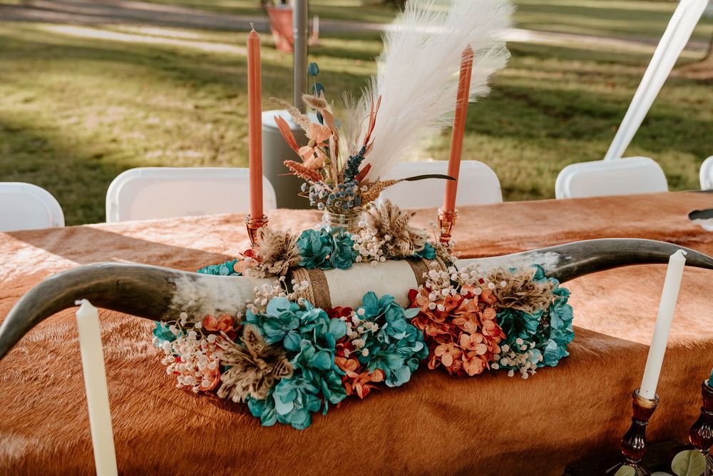 Rustic wedding centerpiece with longhorn, teal flowers, dried botanics, and tall orange candles on cowhide table.