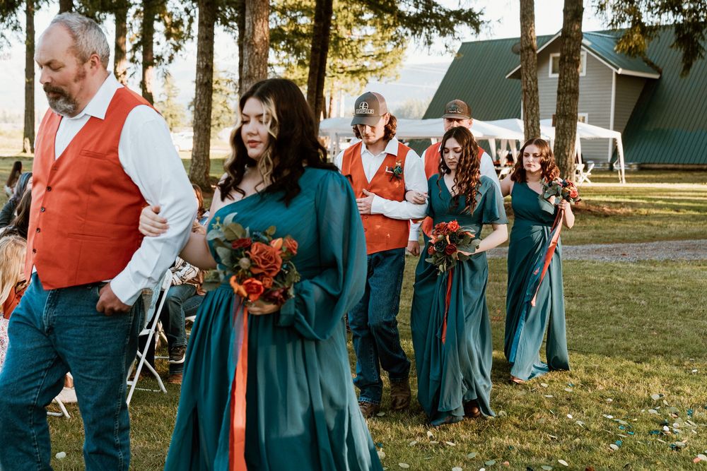 Wedding party in teal dresses and orange vests walk outdoors at a rustic fall wedding ceremony.