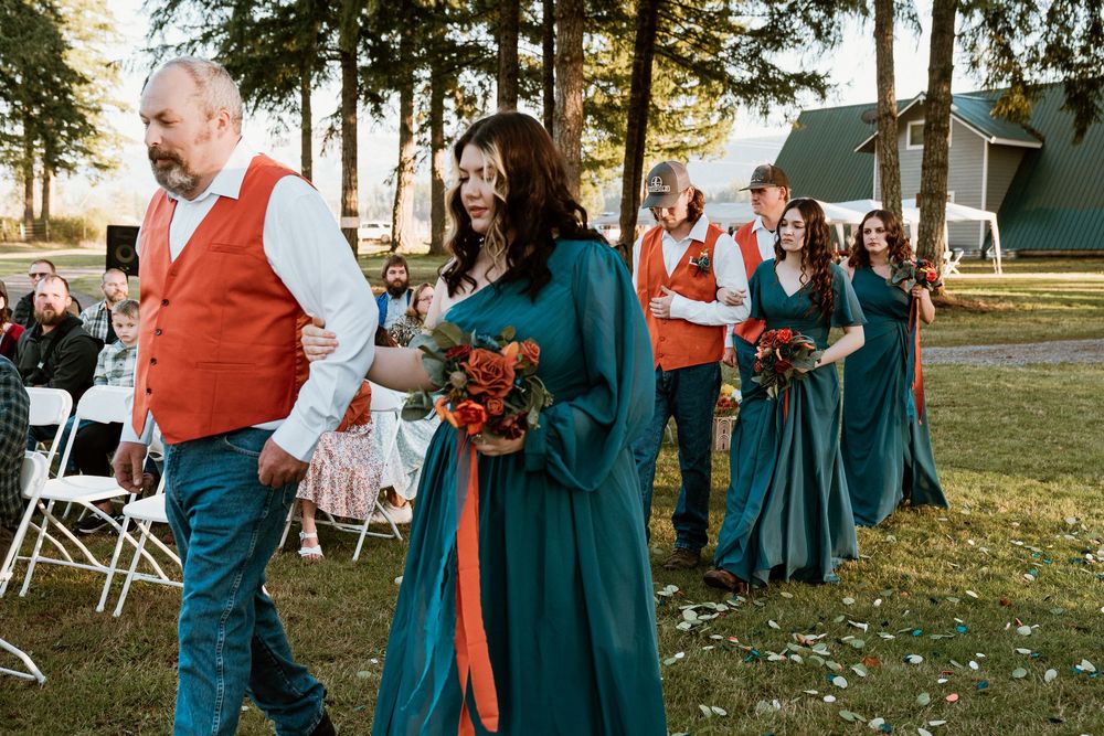 Wedding party in teal gowns and orange vests walking outdoors at a rustic venue ceremony.