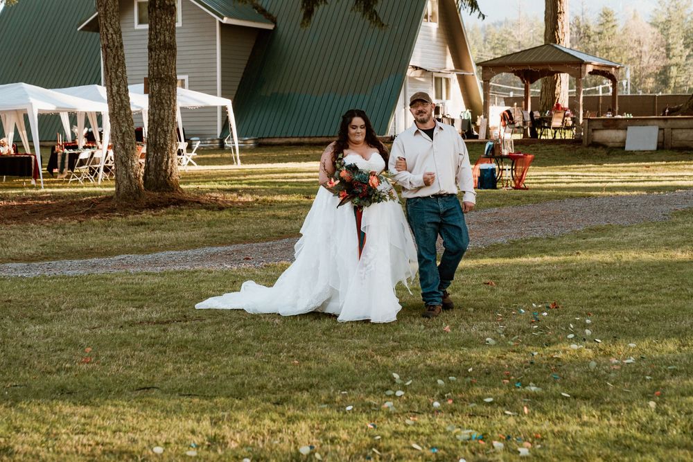 Bride in white gown walks with groom in teal pants outdoors near A-frame chapel on lush green lawn.