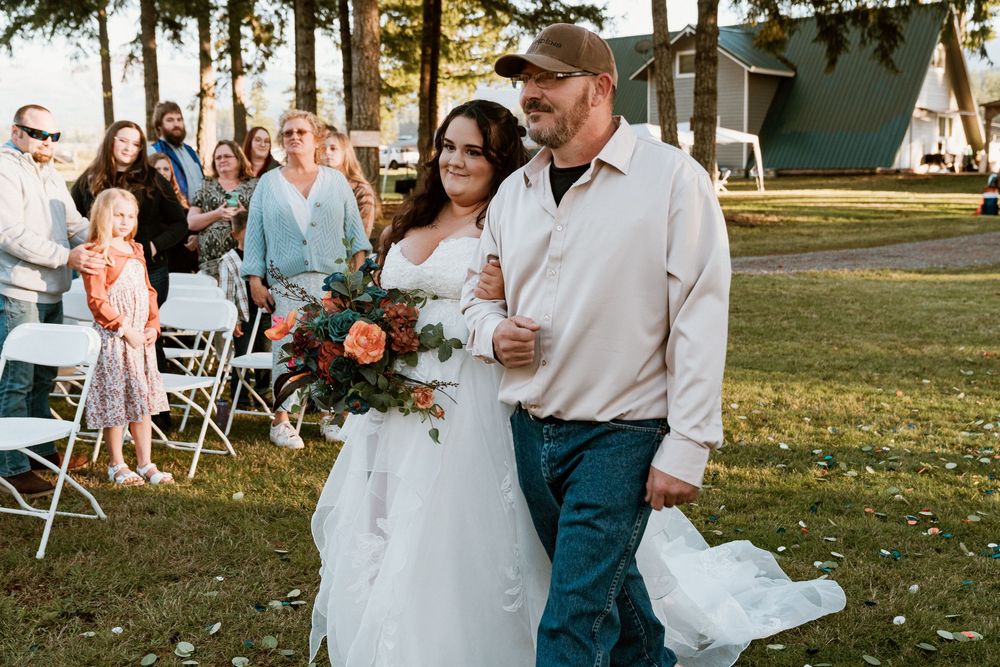 Bride in white gown holding colorful bouquet walks with groom in jeans down outdoor wedding aisle.