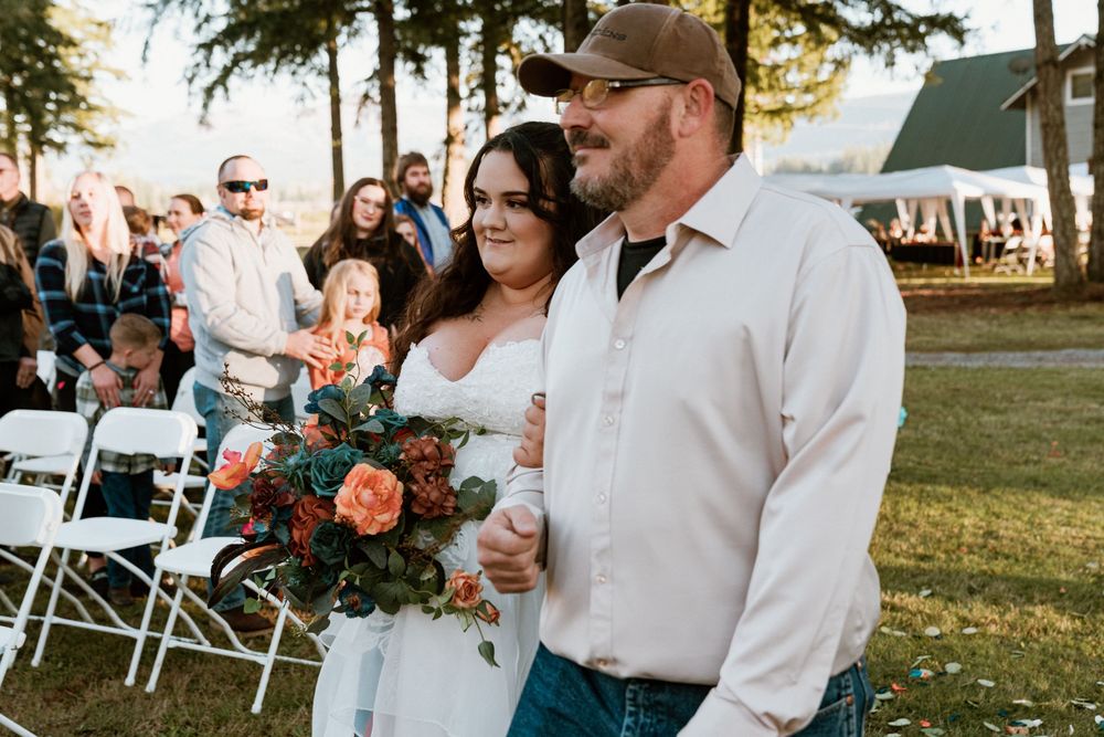 Bride holding colorful bouquet walks down outdoor aisle with escort at rustic wedding ceremony.