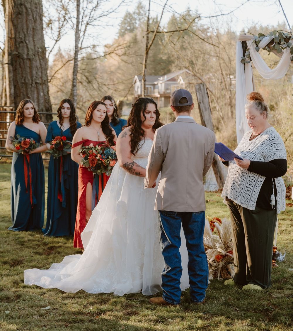 Outdoor wedding ceremony with bride in white gown, bridesmaids in navy dresses, and officiant reading vows in a wooded setting.
