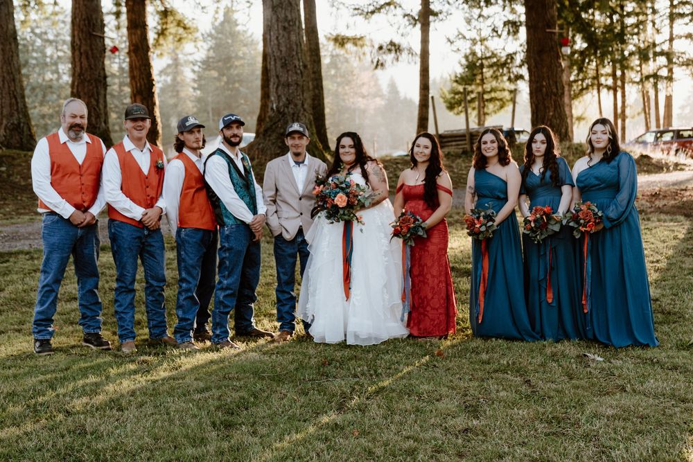Wedding party posing outdoors in forest setting, groomsmen in orange vests with jeans, bridesmaids in teal dresses.