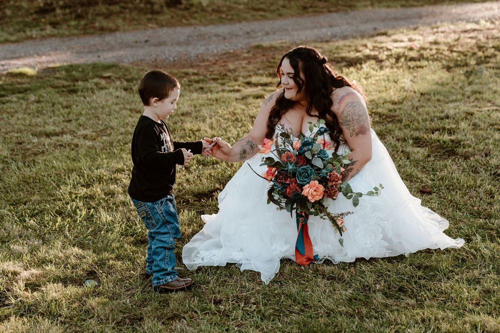 Bride in white gown kneels on grass holding colorful bouquet while interacting with young child in black outfit.