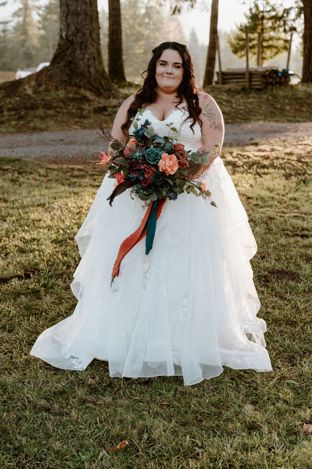 Bride in white ruffled ball gown holding teal and coral bouquet with ribbons, standing outdoors among trees.