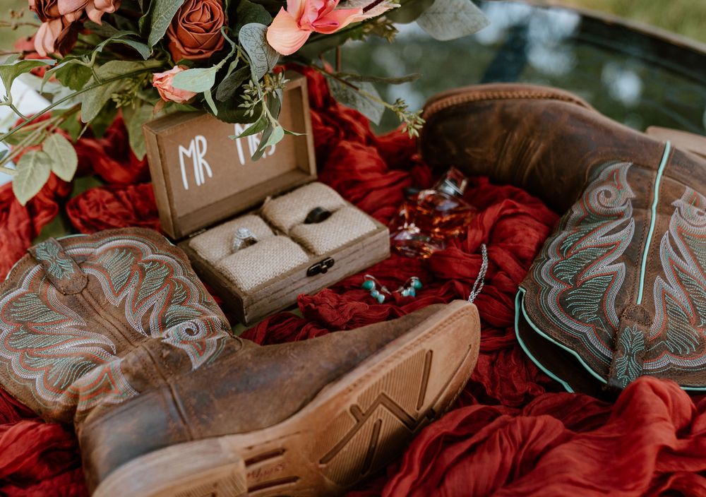Rustic wedding flat lay with cowboy boots, ring box monogrammed MR, wedding bands, and red floral arrangement on crimson fabric.