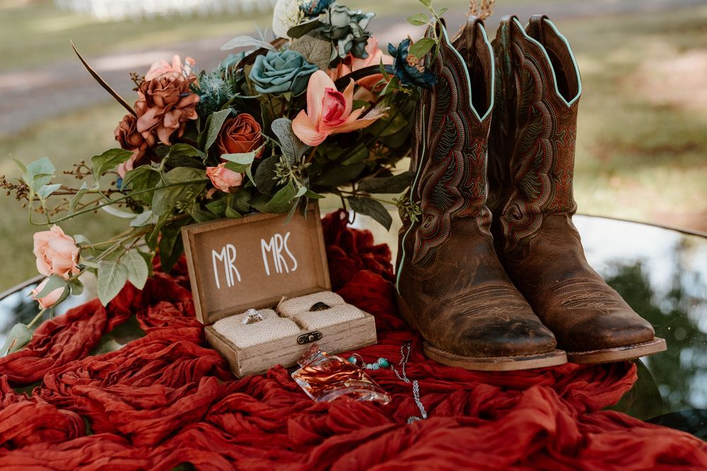 Rustic wedding flat lay with cowboy boots, Mr. & Mrs. ring box, bouquet, and red fabric on a reflective surface.