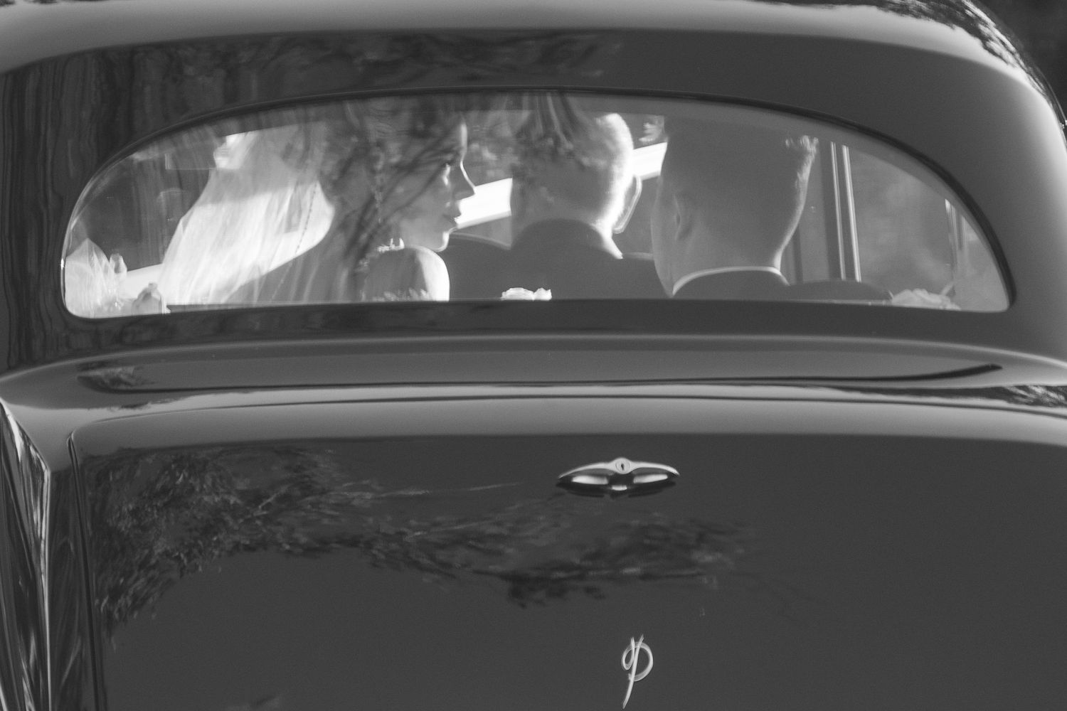 Black and white photo of newlyweds seen through the rear window of a vintage classic car.