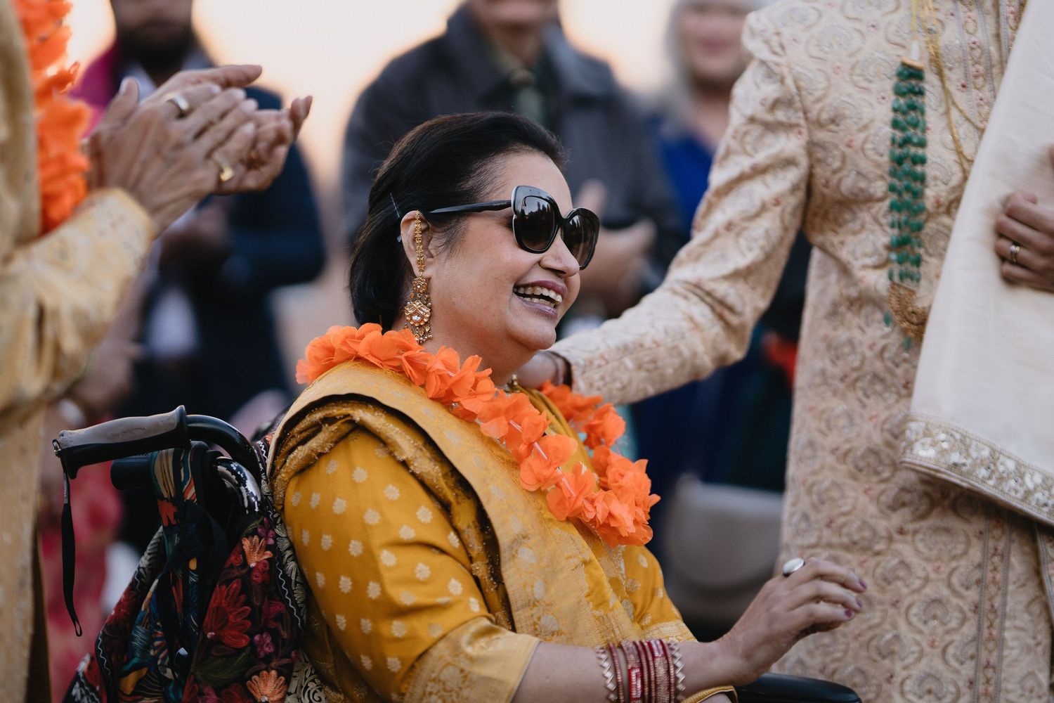 Joyful woman in yellow saree and orange garland smiles while seated in wheelchair at Indian wedding.