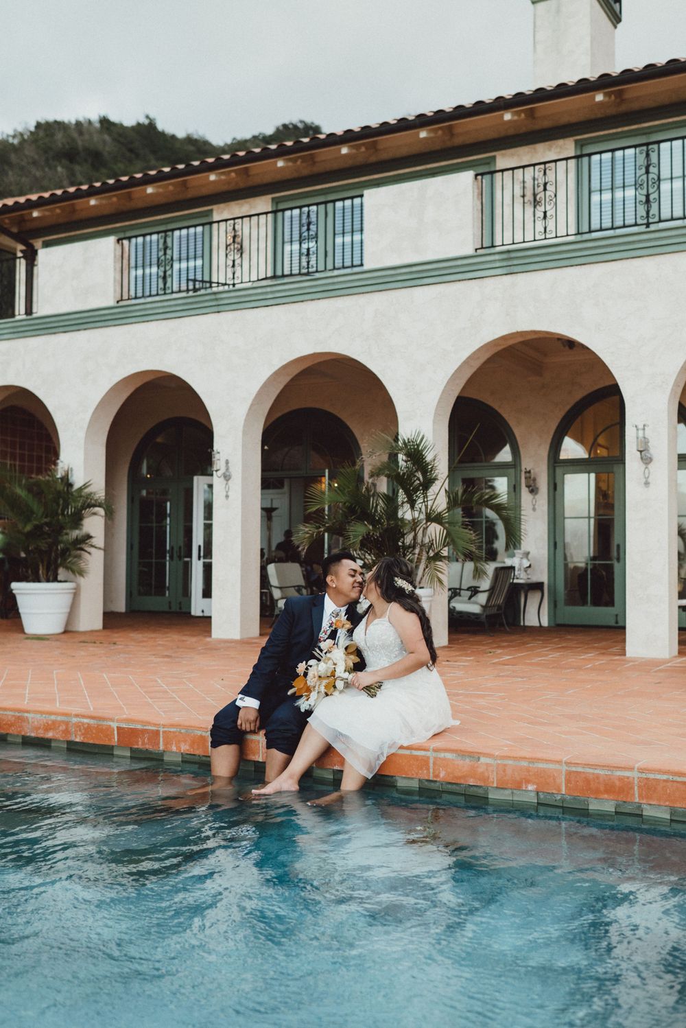 A couple in formal attire playfully falls into a swimming pool at an elegant Spanish-style venue.