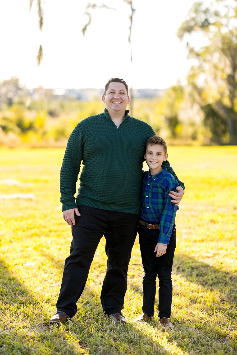 A parent and child stand together outdoors on a sunny day in a grassy field wearing casual clothing.