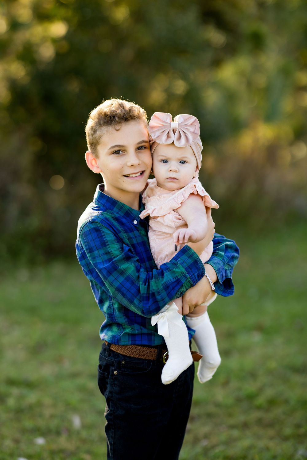A young child in a blue plaid shirt holds a baby wearing pink and white in an outdoor setting with bokeh background.