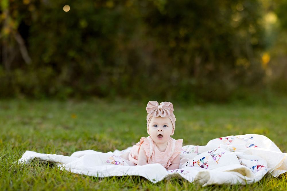 Baby lying on white blanket in grass during outdoor photo session with pink bow accessories.