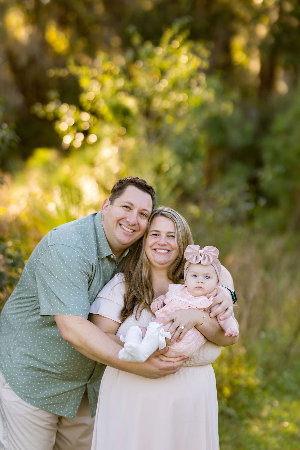 A family portrait taken outdoors in golden sunlight with a baby wearing a pink bow headband.