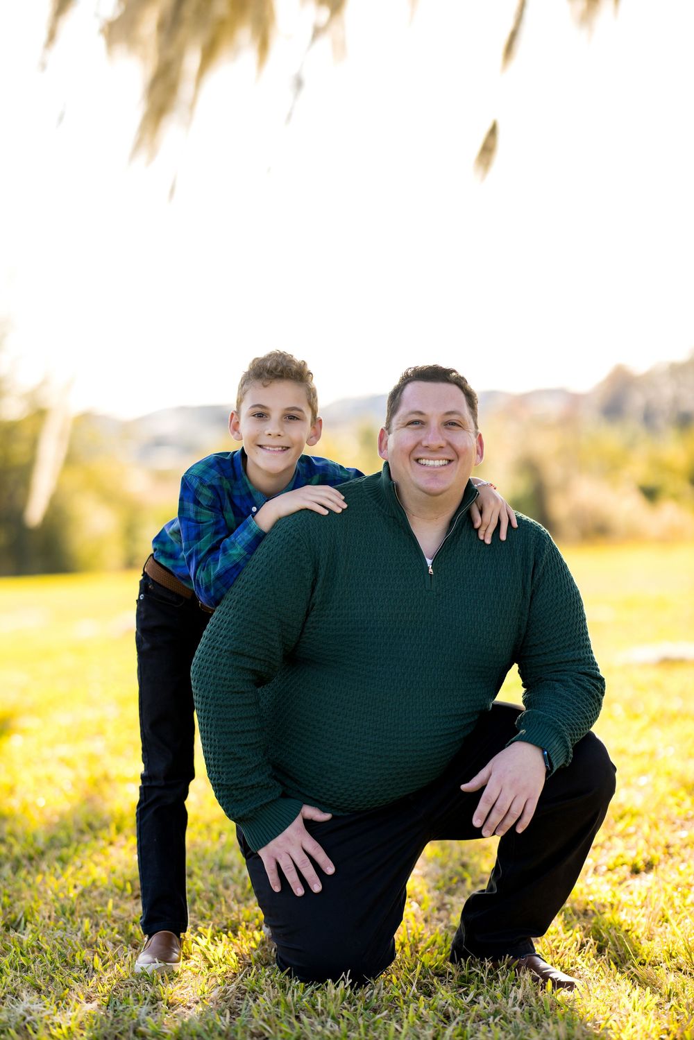 Two people in matching dark sweaters pose together on sunny grass during golden hour.