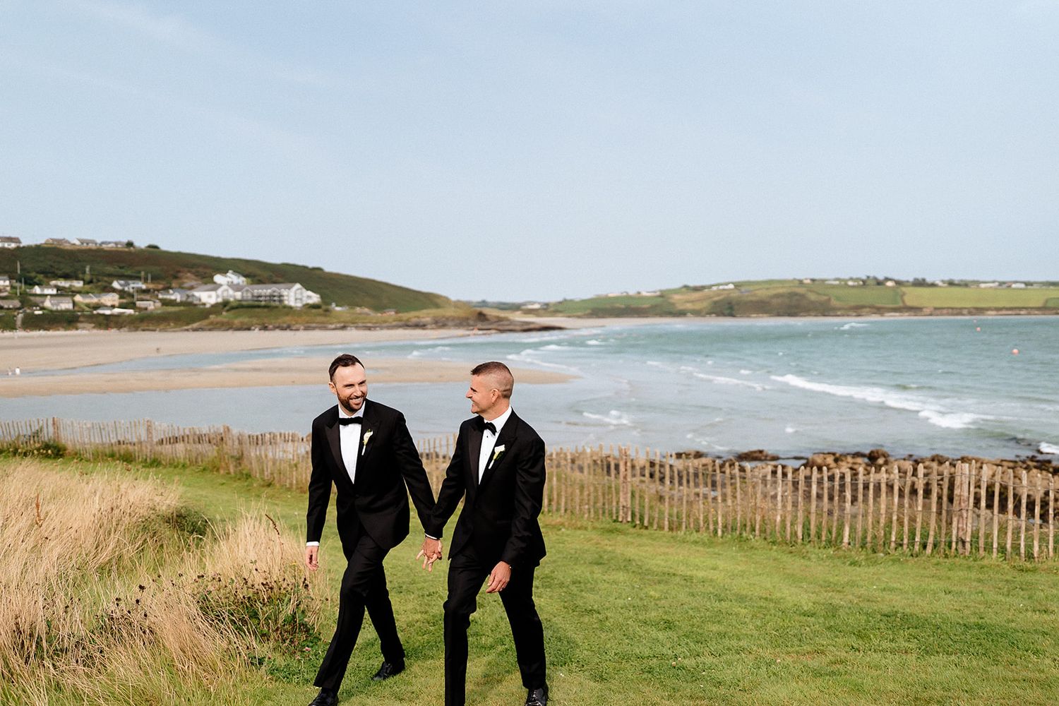 Three people in formal attire walk along a scenic coastal path with wooden fencing and ocean views.
