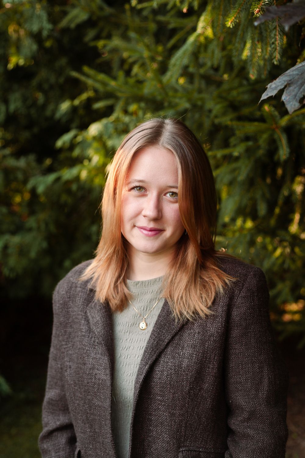 Young woman with reddish-brown hair wearing a gray blazer smiling outdoors near green trees.