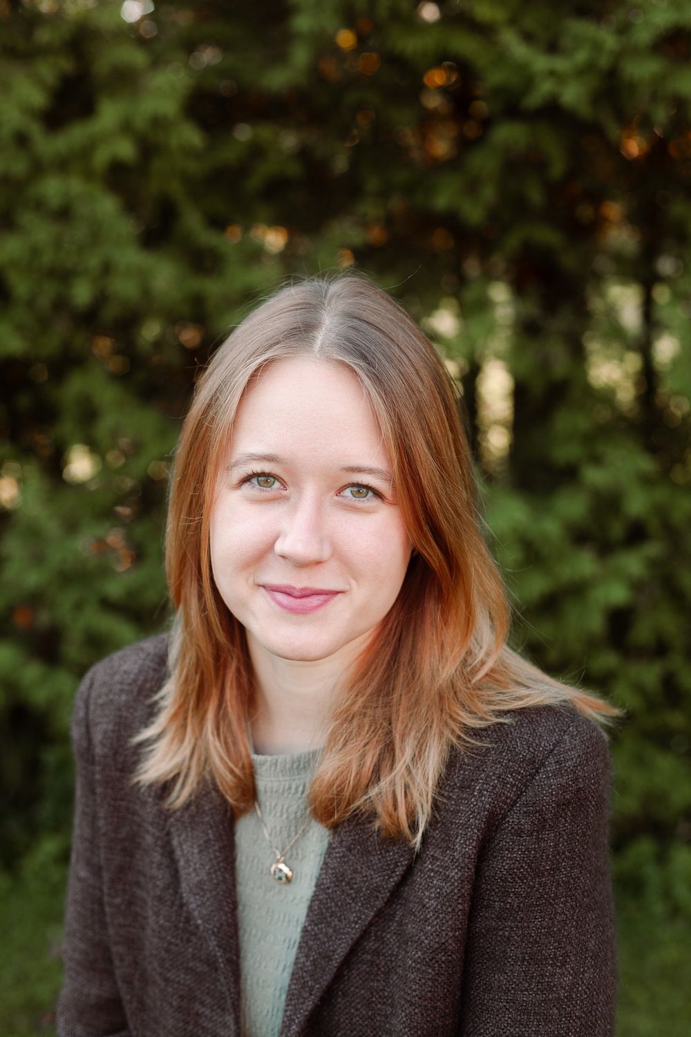 Young woman with auburn hair in a gray blazer smiling warmly in front of green trees.