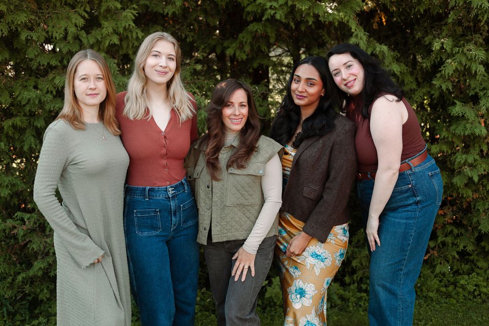 Group of five diverse women smiling together outdoors in front of lush green foliage.