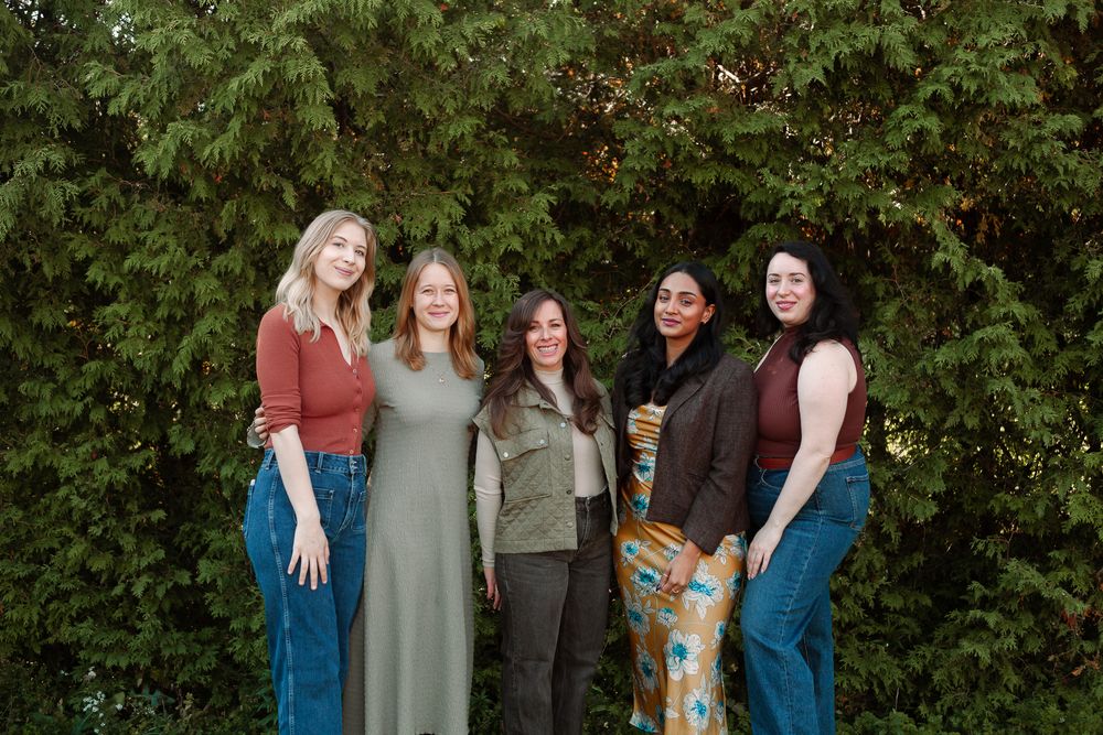 Five women smiling and posing together outdoors in front of lush green trees.
