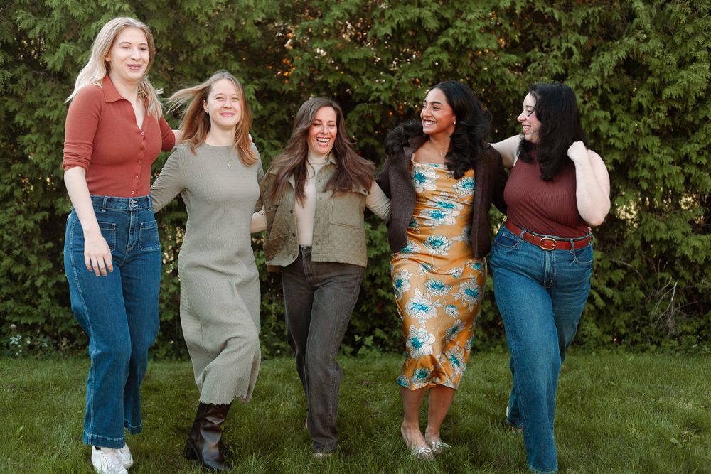 Group of five women laughing and walking together on green grass outdoors.