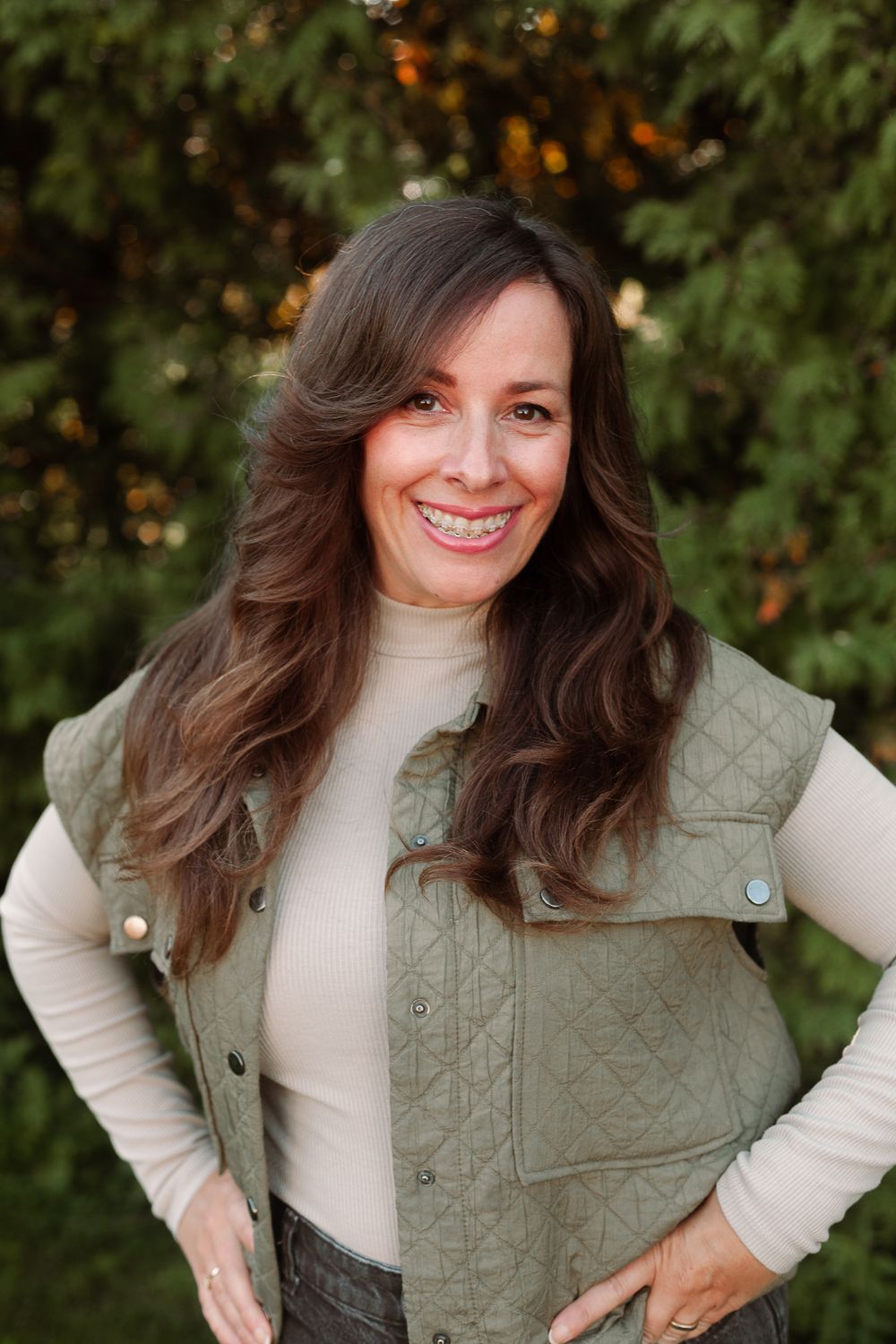 Smiling brunette woman wearing a sage quilted vest over a cream turtleneck outdoors.