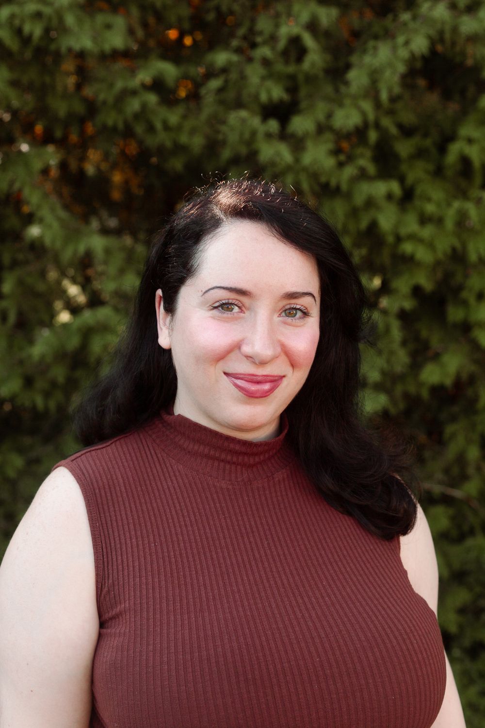 Dark-haired woman in a maroon sleeveless turtleneck smiling softly outdoors.