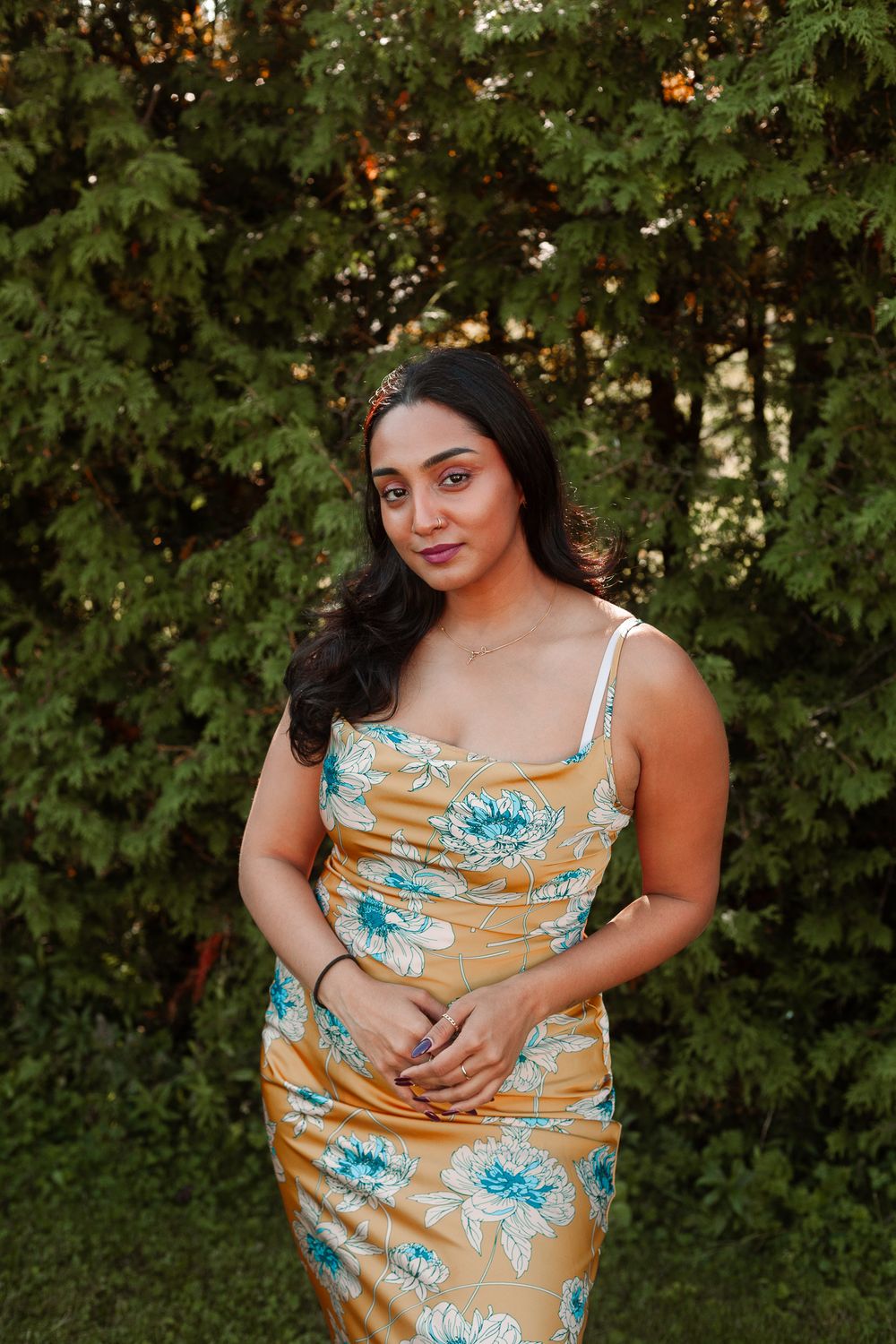 Woman in a gold floral dress posing gracefully in front of green trees outdoors.
